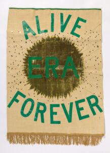 Burlap banner with "ERA Alive Forever" in green tape, stitched on over gold sun and gold sequins. Gold fringe along bottom of banner.