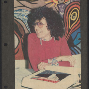 Printed photographic portrait of Judy Chicago sitting in front of colorful artwork with an open book on a table in front of her