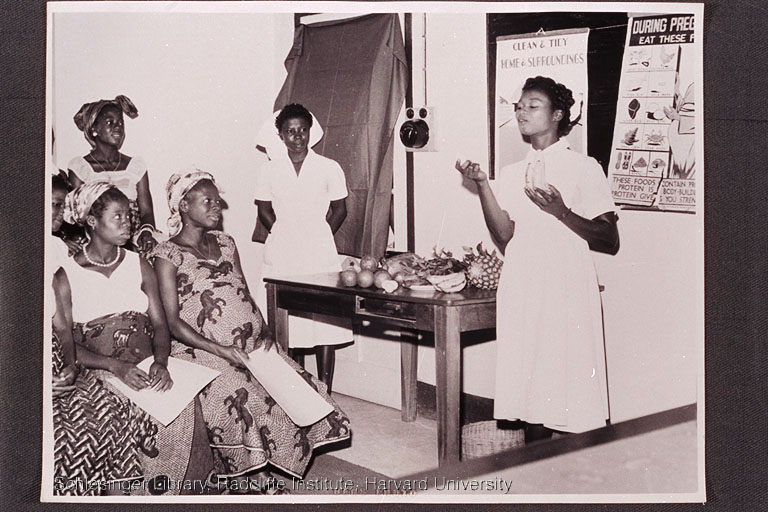  An African nursing student lecturing a group of pregnant women about nutrition while a nurse looks on.