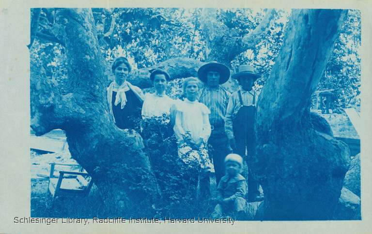 Eve Lummis and her children standing with Native Americans outdoors between trees