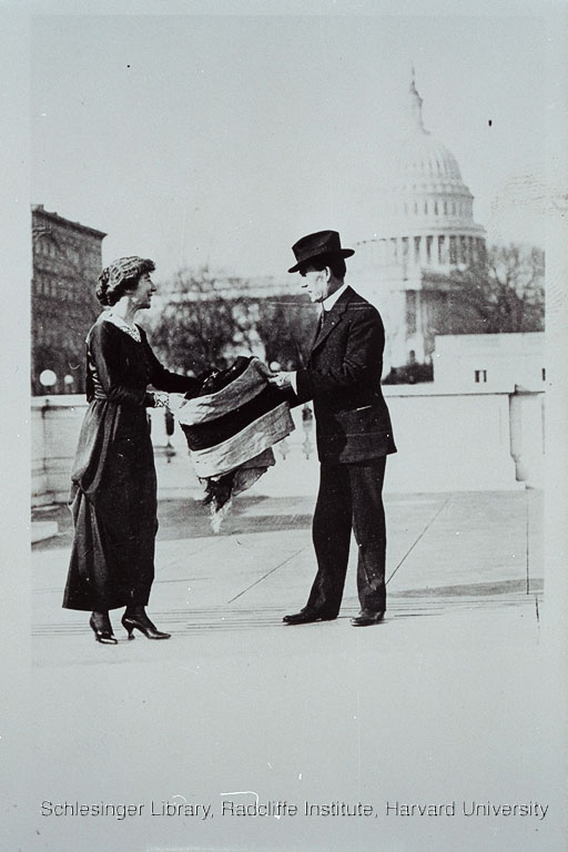 Jeannette Rankin unfurling a flag in front of the Capitol building with a man, probably President Wilson.