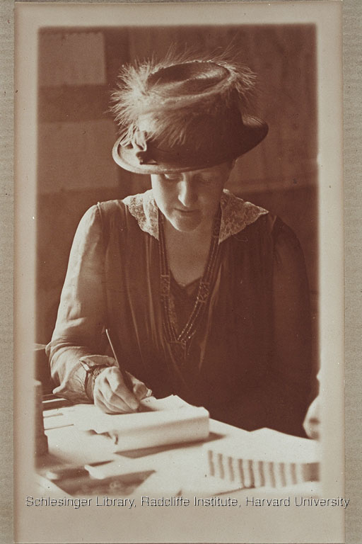 Unidentified woman wearing a feathered hat writing on a notepad at a desk.