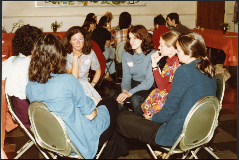 A group of women sit in folding chairs facing each other in a circle as part of a Boston Women's Health Book Collective meeting.