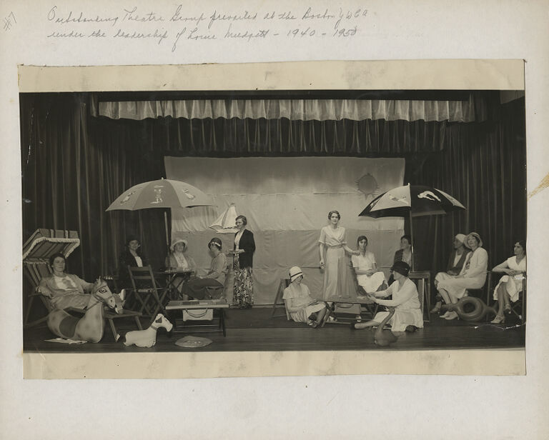 Women sitting on stage in various chairs surrounded by curtains and props.