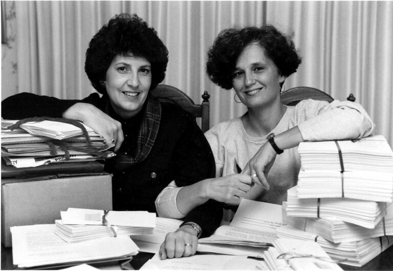 Two women sitting behind a desk stacked with papers, smiling while looking at the camera.