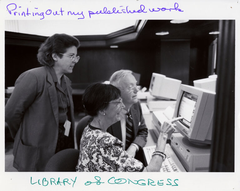 Priscilla Dewey Houghton sitting at a computer and pointing to the screen. The photo is labeled in pen "Printing out my published work. Library of Congress."