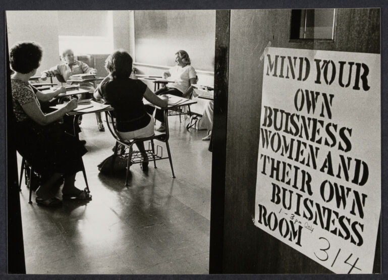 Women sitting in a classroom with a sign on the door that reads "Mind Your Own Business Women and Their Own Business Room"