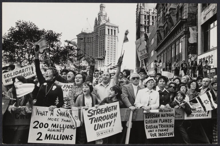 Demonstrators at Soviet Jewish Rally in October, 1973. They are holding signs and cheering. The signs say "Solidarity with Israel," "What a Shame, More than 200 Millions Against 2 Millions," "Strength Through Unity," and "[Obscured] Against Russian Material, Russian Planes, Russian Training. What a Shame for Moscow."