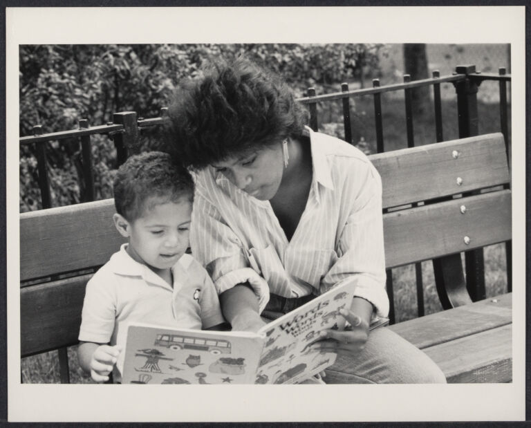 A mother reading a picture book to her child on a park bench.
