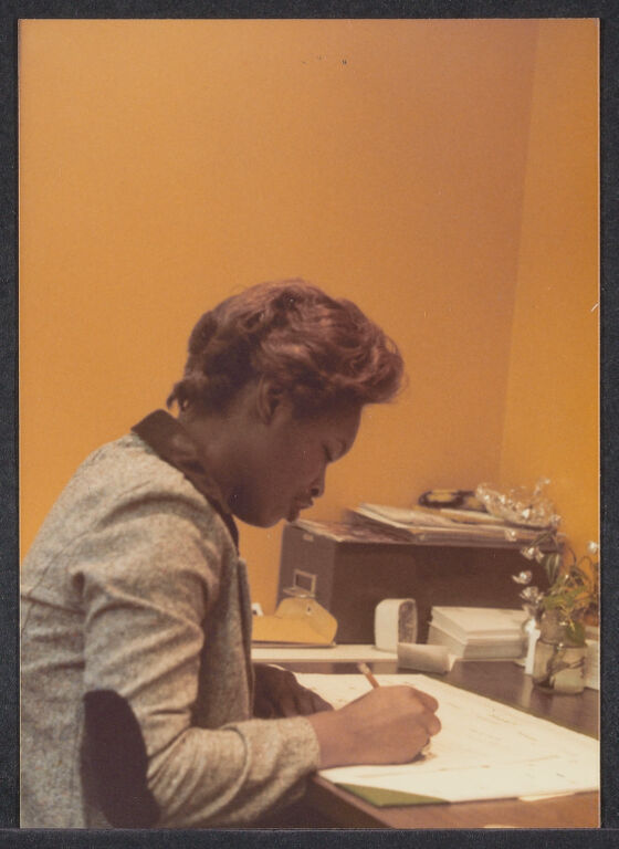 Woman seated at a desk and writing, mostly facing away from camera.